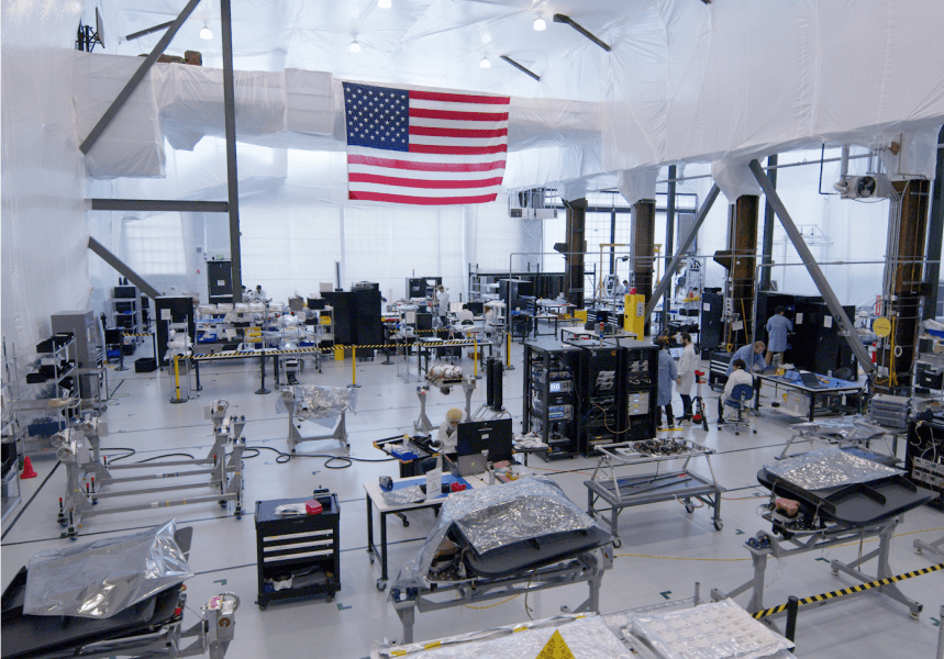 Satellite hardware and workstations inside the Astranis workshop, beneath a large United States flag.